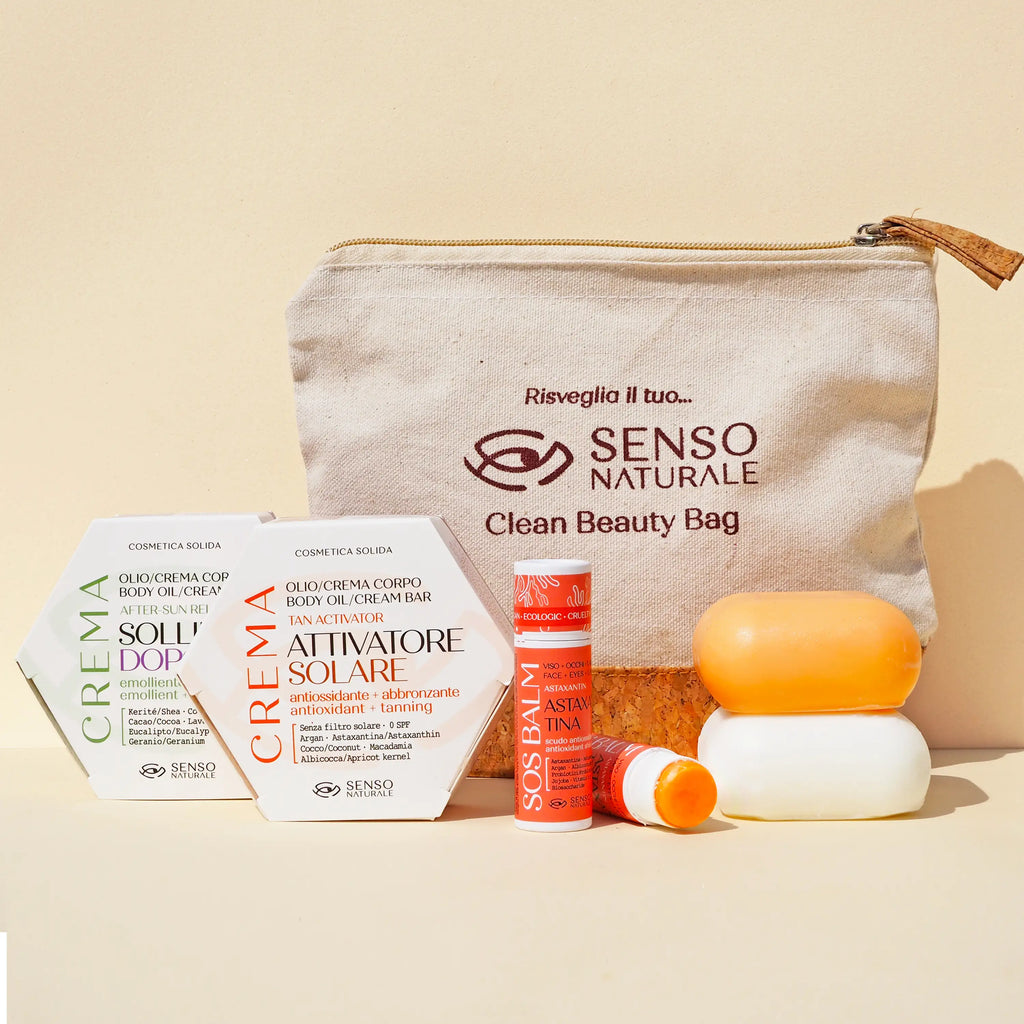 Kit Clean Beauty Bag di Senso Naturale contenente cosmetici solidi: SOS Balm all'astaxantina in stick, Attivatore Solare, Crema Sollievo Doposole e una saponetta arancione. I prodotti sono disposti davanti a una pochette in tessuto naturale color crema su sfondo neutro.