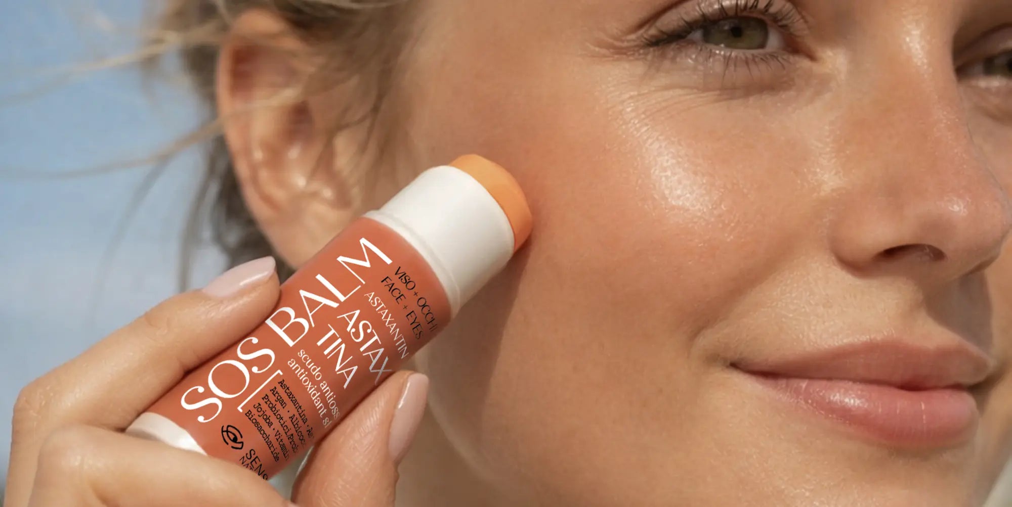 Una donna sorridente applica lo stick SOS Balm all'Astaxantina di Senso Naturale sullo zigomo, mostrando una pelle luminosa, idratata e protetta dal sole.